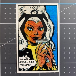 Puede incluir: Una ilustraci&oacute;n de c&oacute;mic de una mujer con cabello blanco y una diadema negra. Lleva una chaqueta negra y una camisa amarilla. Est&aacute; sosteniendo un vaso de l&iacute;quido y mirando al espectador. El texto "I'M NOT BOSSY, I AM THE BOSS" est&aacute; en un globo de di&aacute;logo.