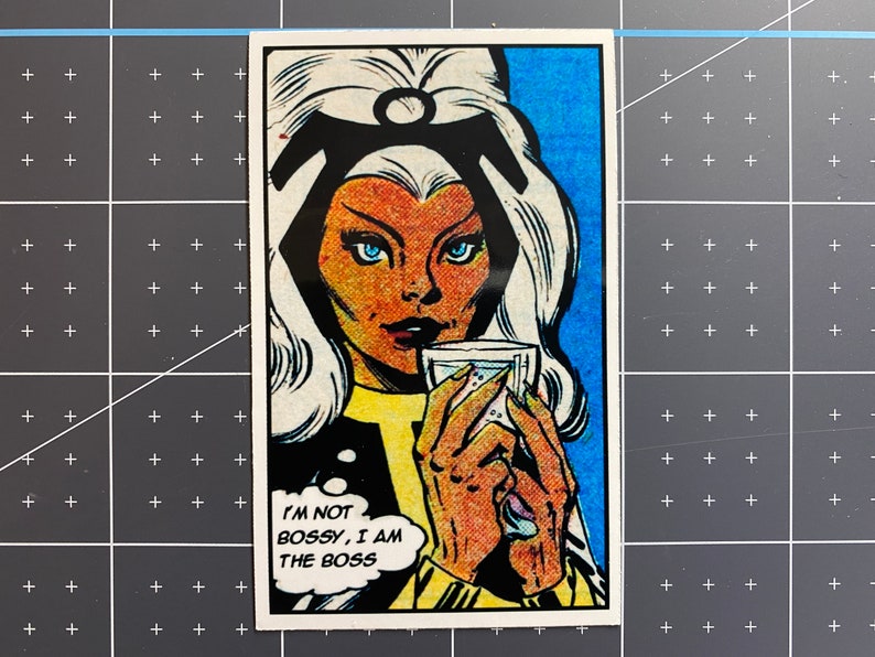 Puede incluir: Una ilustraci&oacute;n de c&oacute;mic de una mujer con cabello blanco y una diadema negra. Lleva una chaqueta negra y una camisa amarilla. Est&aacute; sosteniendo un vaso de l&iacute;quido y mirando al espectador. El texto "I'M NOT BOSSY, I AM THE BOSS" est&aacute; en un globo de di&aacute;logo.