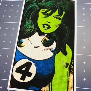 Pu&ograve; includere: Un'illustrazione di fumetto verde e blu di una supereroina con lunghi capelli verdi. La supereroina indossa un costume blu e bianco con un grande numero "4" sul petto.