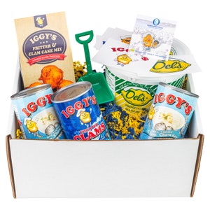 May include: A white gift box filled with various canned goods and a blue and yellow Del's frozen lemonade container. The cans are labeled "IGGY'S" and feature images of clams and clam chowder. The box also includes a bag of IGGY'S Fritter & Clam Cake Mix.