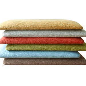 Könnte beinhalten: Sechs unterschiedlich farbige Kissen, die übereinander gestapelt sind. Die Kissen bestehen aus einem weichen, strukturierten Stoff. Die Farben sind gelb, grau, rot, grün, blau und braun.