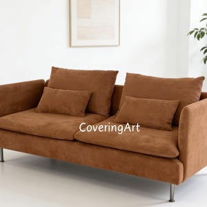 May include: A brown two-seater sofa with two large back cushions and two smaller rectangular cushions. The sofa has a low profile, with a simple design and metal legs. The text "CoveringArt" is visible on the sofa.