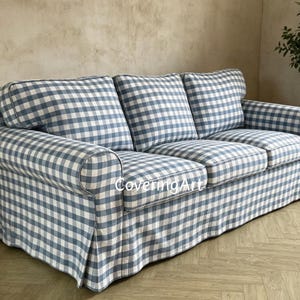 May include: A three-seater sofa upholstered in blue and white gingham fabric. The sofa features rolled arms and a skirted base. The fabric has a light blue and white check pattern. The sofa is in a room with a light-colored wall and a wooden floor.