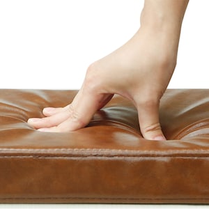 Puede incluir: Primer plano de un cojín de cuero marrón con una mano presionando el centro. El cojín está acolchado y tiene un borde cosido.