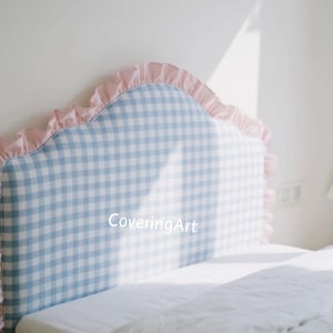 Può includere: Una testiera a quadretti blu e bianchi con un bordo rosa a balze. La testiera è posta sopra un letto bianco con una trapunta bianca. Il testo "CoveringArt" è visibile.