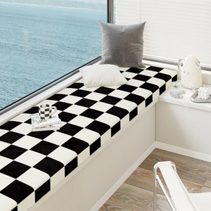 Puede incluir: Un cojín para asiento de ventana con un patrón de tablero de ajedrez en blanco y negro. El cojín es rectangular y se asienta sobre una base blanca. Dos almohadas, una gris y otra blanca, descansan sobre el cojín. Una taza en blanco y negro y libros están sobre el cojín.
