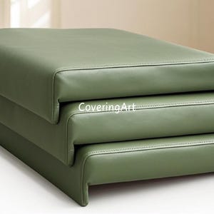May include: A stack of three green leather cushions. The cushions are rectangular and have a smooth, soft surface. The top cushion has the text "CoveringArt" printed on it.