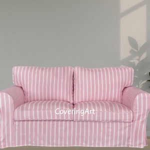Op de afbeelding: Een roze en wit gestreepte bank met twee zitkussens en twee rugkussens. De bank heeft gerolde armen en een rokbasis. De stof heeft verticale strepen. De bank staat voor een lichtgrijze muur.