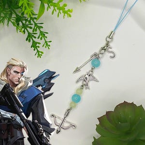 May include: A silver charm with a light blue cord, featuring a bow and arrow, a bird, and a small arrow. The charm has light blue and yellow beads. The background includes a green plant and a cartoon character.