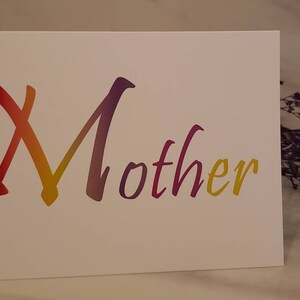 May include: A white greeting card with the word "Mother" written in a colorful, gradient font. The colors transition from red to yellow to purple. A sprig of dried purple flowers is in the background.