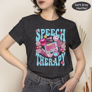 May include: Dark grey heather t-shirt featuring a retro-style graphic that reads "Speech Therapy" in blue and pink lettering. The graphic includes a pink mouth, a smiley face, a keyboard, and other colourful illustrations.