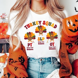 May include: White t-shirt with a colorful "TURKEY SQUAD" graphic featuring turkey illustrations and the acronyms PT, SLP, and OT. The shirt has the phrases "Helps That Hobble", "Helps You Gobble", and "Helps That Wobble". A pumpkin-shaped mug is held in the image.