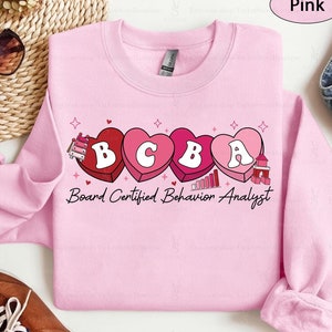 May include: Pink sweatshirt with a graphic of hearts spelling out "BCBA" with the text "Board Certified Behaviour Analyst" below.