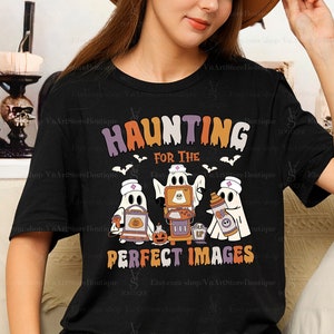 May include: Black t-shirt with the text "HAUNTING FOR THE PERFECT IMAGES" in orange, purple, and white. The shirt features cartoon ghosts with medical symbols and Halloween-themed props, including a pumpkin and bats.