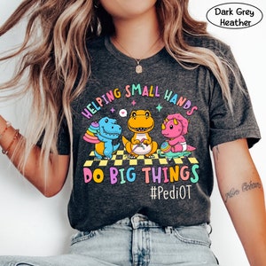 May include: Dark grey heather t-shirt with a graphic of three dinosaurs. The dinosaurs are holding toys and the text "Helping Small Hands Do Big Things #PediOT" is printed on the shirt.