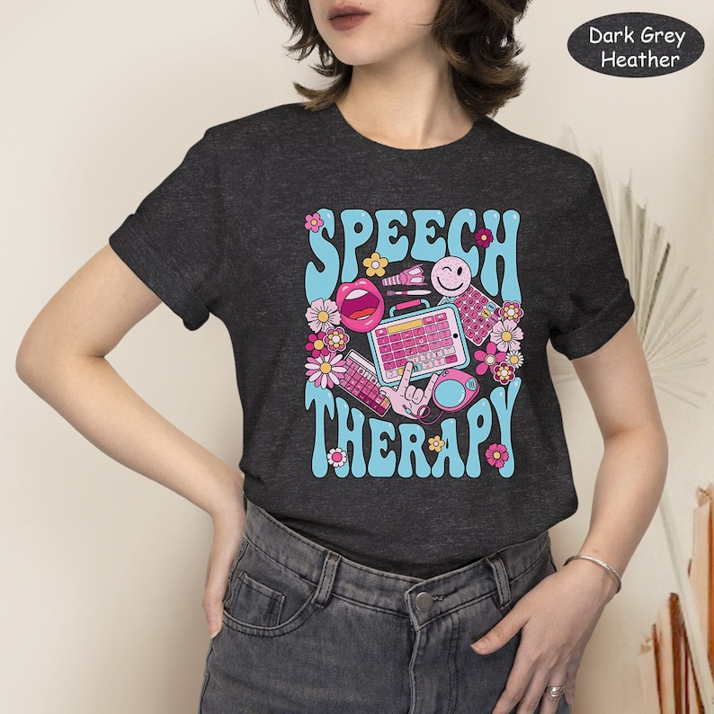 May include: Dark grey heather t-shirt featuring a retro-style graphic that reads "Speech Therapy" in blue and pink lettering. The graphic includes a pink mouth, a smiley face, a keyboard, and other colourful illustrations.