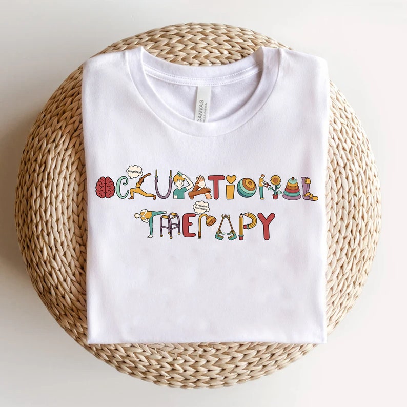 May include: White t-shirt with the words "Occupational Therapy" spelled out in colourful letters. Each letter is decorated with an image related to occupational therapy, such as a brain, a sunflower, a pencil, and a walker.