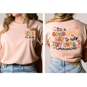 Op de afbeelding: Een roze T-shirt met een kleurrijke afbeelding die op de achterkant "It's a good day to help tiny humans communicate" en op de voorkant "SLP Mr. Brown" zegt.