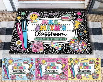 May include: A black and white composition notebook-style doormat with the text "MRS. SMITH'S Classroom" in colorful letters. The mat includes various sticker-like graphics, such as pens, a smiley face, and an apple. The text "I'm so happy to see you here" is also included.