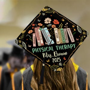 May include: Black graduation cap decorated with a floral design and books with the words "Balance", "Endurance", "Mobility", "Strength", "ADLs", "Coordination", "Safety", "Prevention", and "Performance". The text "Physical Therapy Mrs. Brown 2025" is also on the cap.