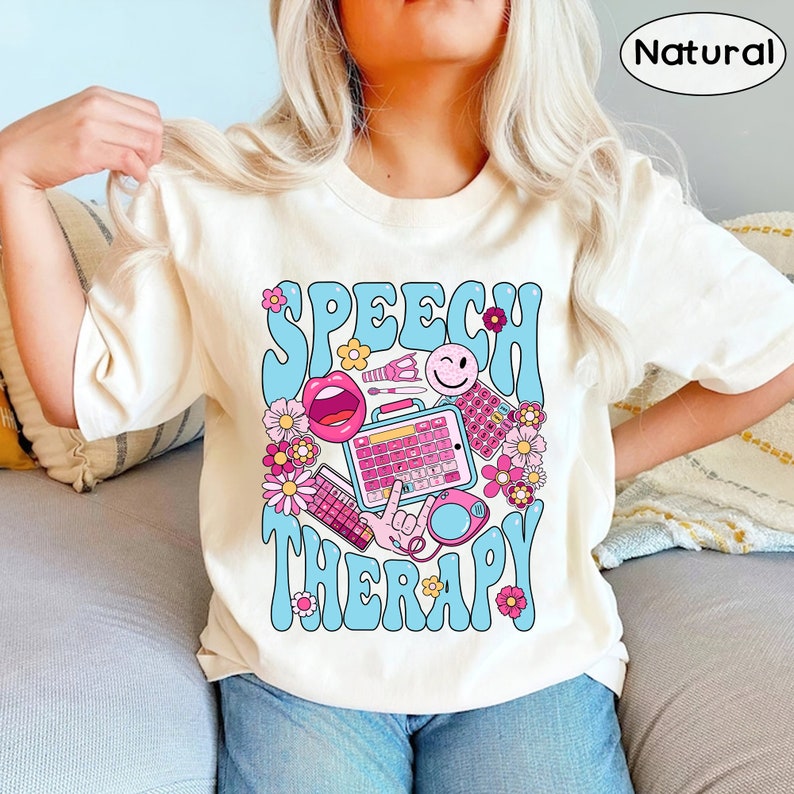 May include: A white t-shirt featuring a blue and pink graphic design that reads "Speech Therapy". The design includes a speech bubble, a microphone, a smiley face, and other speech therapy related items.