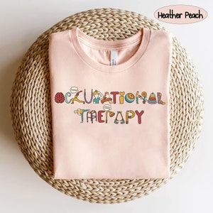 May include: A pink t-shirt with the words "Occupational Therapy" spelled out in colourful letters. Each letter is decorated with a small illustration related to occupational therapy, such as a brain, a hand, and a walker.