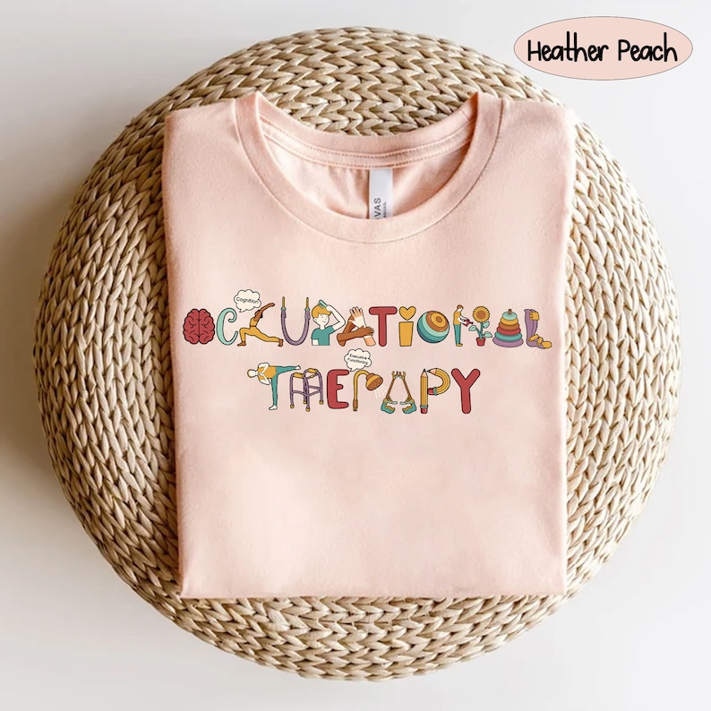 May include: A pink t-shirt with the words "Occupational Therapy" spelled out in colourful letters. Each letter is decorated with a small illustration related to occupational therapy, such as a brain, a hand, and a walker.