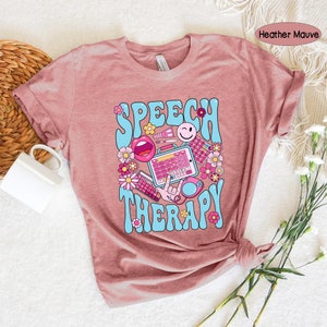 May include: A heather mauve t-shirt featuring a graphic design of the words "Speech Therapy" in a retro style with colourful illustrations of speech therapy tools and flowers.