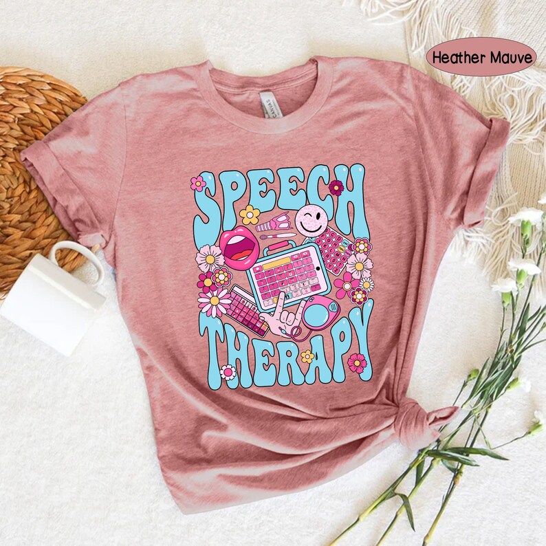 May include: A heather mauve t-shirt featuring a graphic design of the words "Speech Therapy" in a retro style with colourful illustrations of speech therapy tools and flowers.