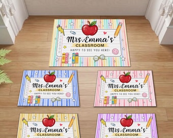 Op de afbeelding: Vijf rechthoekige deurmatten met een gelinieerd papierontwerp en verticale pastelstrepen. Elke mat toont een rode appel, een geel potlood, een paperclip, een bril en de tekst Mrs. Emma's CLASSROOM HAPPY TO SEE YOU HERE in het zwart.