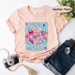 May include: A peach-coloured t-shirt with the text "SPEECH THERAPY" in blue and pink letters. The text is surrounded by colourful illustrations of speech therapy tools, including a mouth, a keyboard, and a microphone.