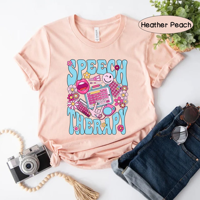 May include: A peach-coloured t-shirt with the text "SPEECH THERAPY" in blue and pink letters. The text is surrounded by colourful illustrations of speech therapy tools, including a mouth, a keyboard, and a microphone.