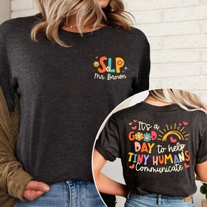 Op de afbeelding: Een donkergrijs T-shirt met de tekst "SLP" in kleurrijke letters met bloemen en glitter. De tekst "Mr. Brown" staat onder de letters. De achterkant van het shirt heeft de tekst "It's a good day to help tiny humans communicate" in kleurrijke letters met bloemen en een zon.