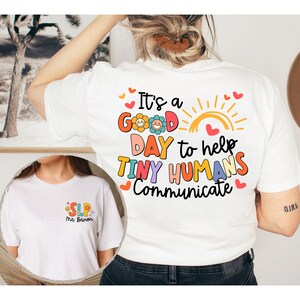 Op de afbeelding: Wit T-shirt met de tekst "It's a good day to help tiny humans communicate" in kleurrijke letters met een zon- en bloemenmotief. De voorkant van het shirt heeft de tekst "SLP" en "Mrs. Brown" in een kleurrijk lettertype.