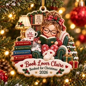 May include: A decorative Christmas ornament featuring a girl with glasses reading a book in a green armchair with a cat. The ornament includes a stack of books, a lamp, a small tree, and text that reads: Book Lover Claire, Booked for Christmas 2026.