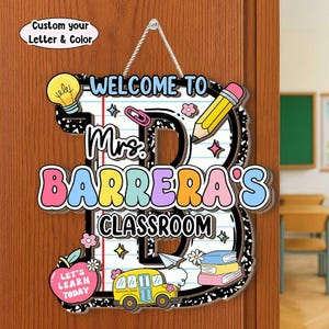 May include: A hanging classroom door sign shaped like the letter B with a notebook paper background. It features the text "WELCOME TO Mrs. BARRERA'S CLASSROOM" and "LET'S LEARN TODAY" with illustrations of a lightbulb, pencil, bus, and books.