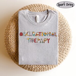 May include: A heather grey t-shirt with the words "Occupational Therapy" spelled out in colourful letters. Each letter is decorated with an image related to occupational therapy, such as a brain, a hand, a walker, and a ball.