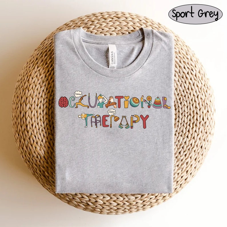 May include: A heather grey t-shirt with the words "Occupational Therapy" spelled out in colourful letters. Each letter is decorated with an image related to occupational therapy, such as a brain, a hand, a walker, and a ball.