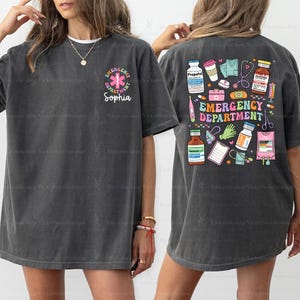 May include: Two gray t-shirts with a colorful graphic design of medical supplies and the text "Emergency Department". One shirt has the text "Emergency Department" with a pink star of life and the name "Sophia" underneath.