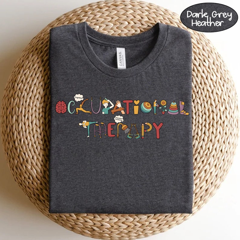May include: Dark grey heather t-shirt featuring the text "Occupational Therapy" in colourful letters. Each letter is decorated with an illustration related to occupational therapy, such as a brain, a hand, a walker, and a ball.