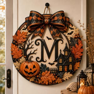 May include: A circular Halloween wall hanging featuring a large orange jack-o'-lantern, a black haunted house, and a central letter M. The piece is decorated with orange and black autumn leaves, small black bats, and a large plaid bow at the top.