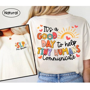 Op de afbeelding: Een wit T-shirt met de tekst "It's a good day to help tiny humans communicate" in kleurrijke letters met een zon en bloemen. Het shirt heeft ook kleine hartjes rond de tekst.