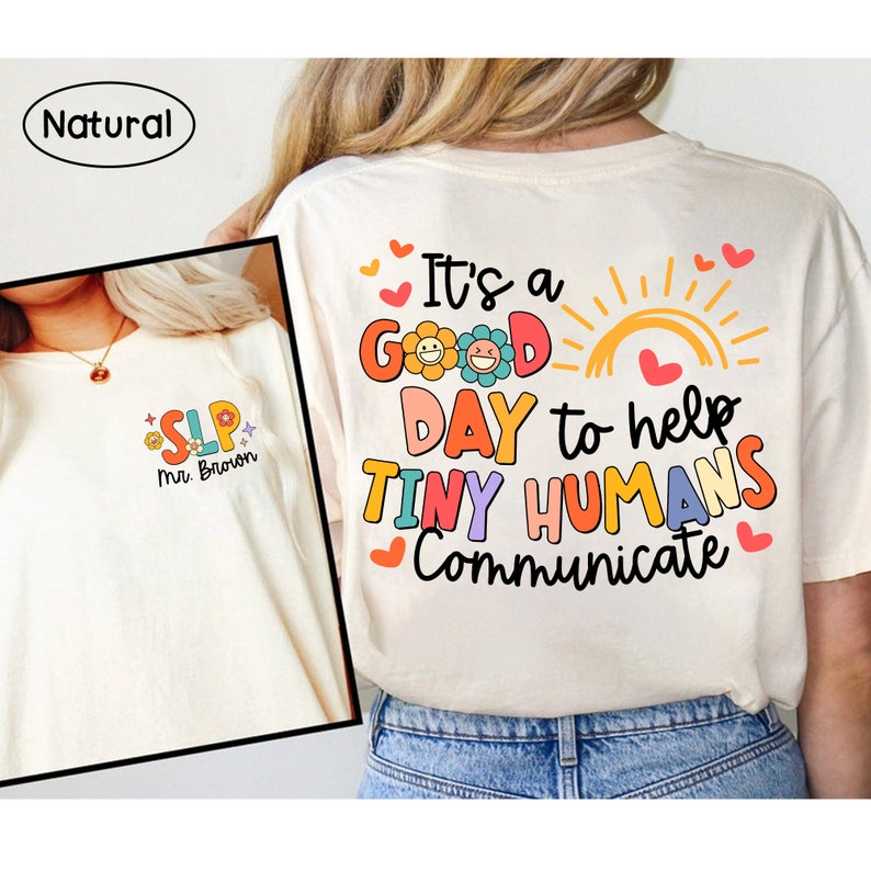 Op de afbeelding: Een wit T-shirt met de tekst "It's a good day to help tiny humans communicate" in kleurrijke letters met een zon en bloemen. Het shirt heeft ook kleine hartjes rond de tekst.