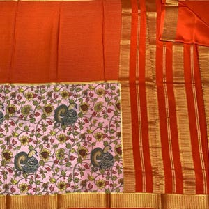 May include: A vibrant orange saree featuring a textured grid pattern on the main body, a wide border with vertical gold stripes, and a contrasting light pink panel decorated with a repeating pattern of dark grey peacocks and yellow flowers.