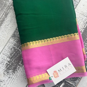 May include: A folded textile featuring a deep green upper section and a vibrant pink lower section. A gold-colored border with a repeating design separates the two colors. A white tag with the text "MIRA SILKS" is attached.