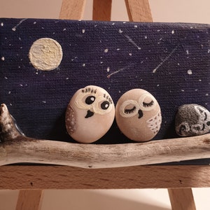 May include: A small canvas painting on an easel depicts two white owls perched on a branch under a starry night sky. The owls have painted eyes and beaks, and the moon is visible in the background.