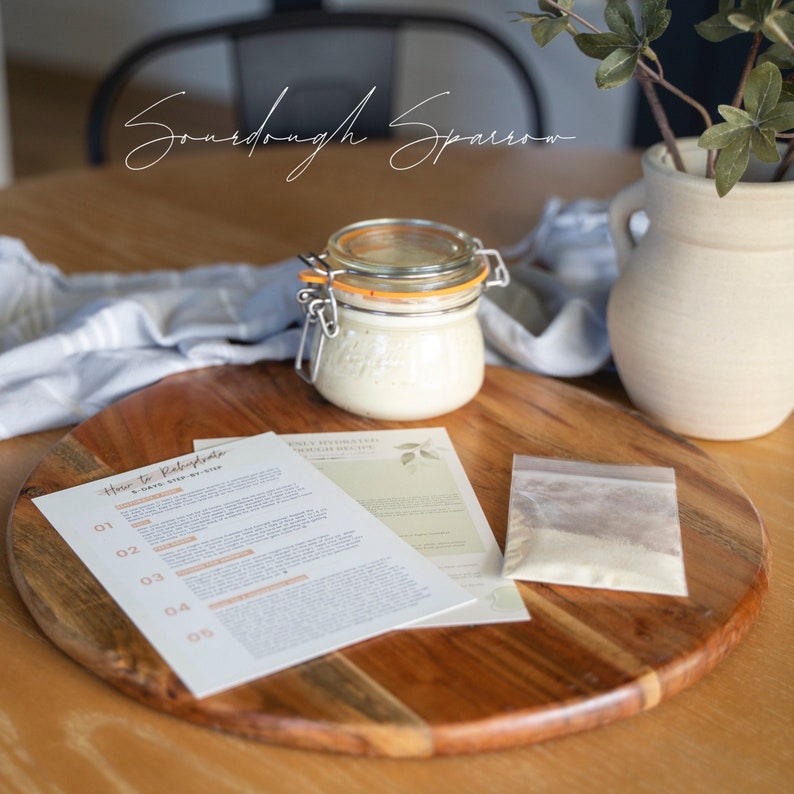 May include: A sourdough starter kit with a glass jar of starter, a packet of sourdough flour, and instructions on how to make sourdough bread. The kit is on a wooden cutting board.