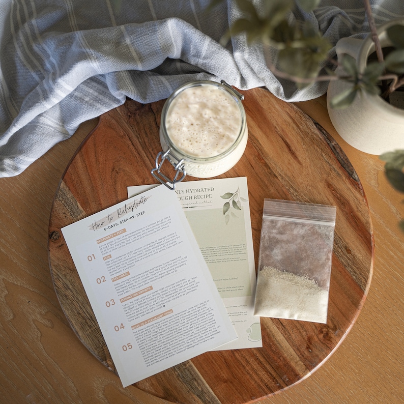 May include: A close-up of a wooden cutting board with a glass jar of sourdough starter, a small plastic bag of sourdough starter, and a printed instruction sheet titled "How to Rehydrate 5-Days: Step-by-Step" and "Only Hydrated Dough Recipe".