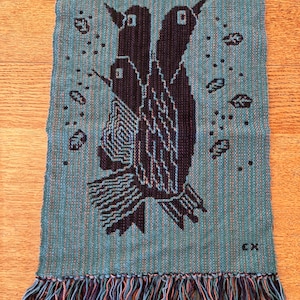 May include: A teal woven textile featuring a black bird design with three birds and leaf accents. The textile has a fringed bottom with multicolored threads. The initials "CX" are in the bottom right corner.