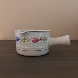 Könnte beinhalten: Weißer Keramik-Soßenbehälter mit Griff und Ausguss. Das Gefäß ist mit einem Blumenmuster in Blau, Rosa und Grün verziert. Eine dünne kastanienbraune Linie akzentuiert den Rand und die Basis. Der Soßenbehälter steht auf einer dunkelbraunen Oberfläche.